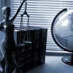 A state of Lady Justice holding the scales of justice standing next to legal books and a globe.
