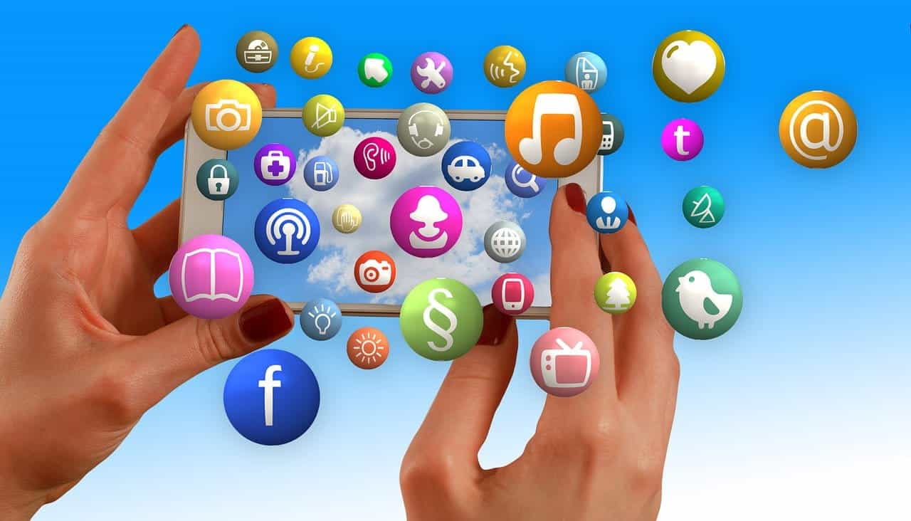 Sepasang tangan memegang smartphone secara horizontal sementara banyak ikon berbeda untuk platform dan aplikasi keluar dari layarnya.