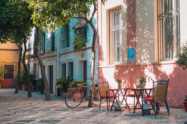 A colorful street in Spain with tables outside in sunlight and trees.