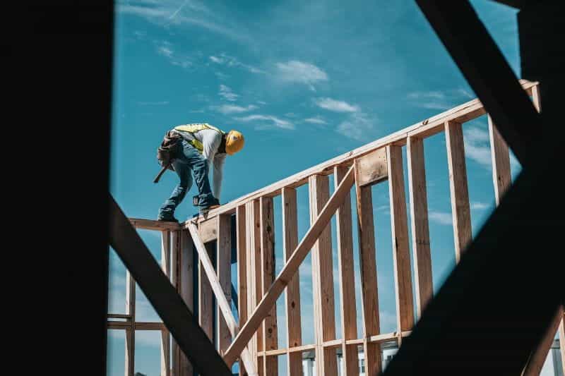 Seorang pekerja konstruksi dengan topi keras bekerja di atas kerangka bangunan kayu, dengan langit biru di belakangnya.