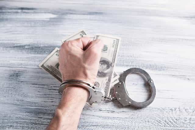 A hand in a handcuff is gripped over a table laden with cash.