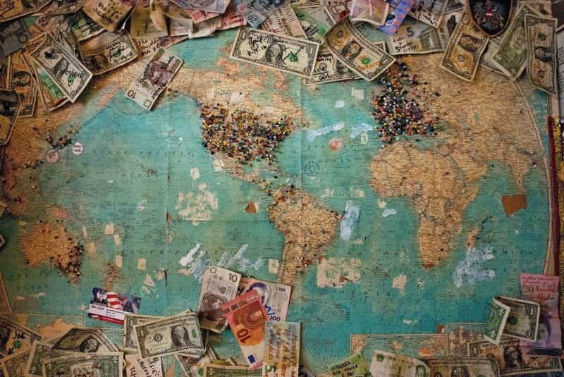 An antique world map displays various currencies and push pins all over it.