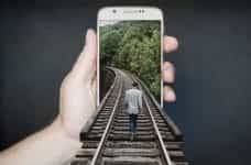 A person holding up a smartphone which has a man walking along train tracks emerging out of it.