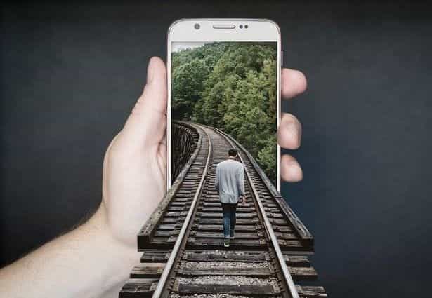 Seseorang yang memegang smartphone yang memiliki seorang pria berjalan di sepanjang rel kereta api muncul dari sana.