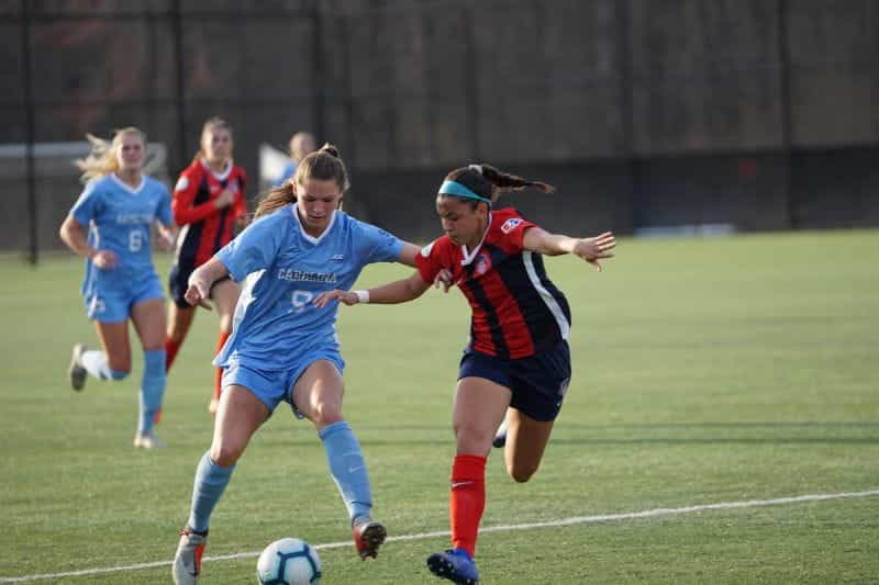 Sekelompok wanita bermain sepak bola dengan kaus biru dan merah.