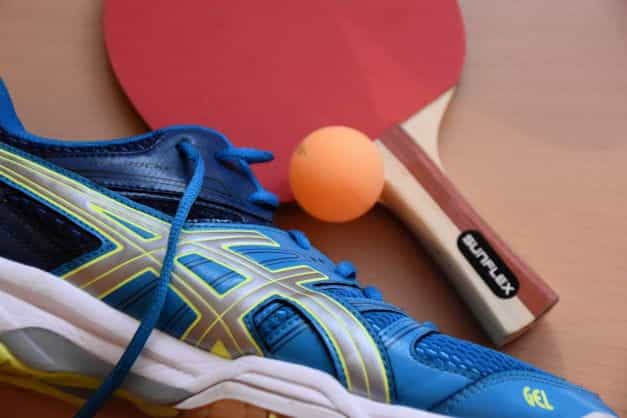 Sepatu olahraga biru duduk di sebelah bola tenis meja dan raket.