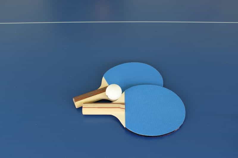 Dua raket tenis meja tergeletak berdampingan di lantai dengan bola tenis meja di tengahnya.