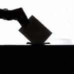 A silhouette of a hand dropping a ballot into a ballot box.