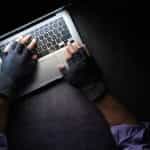Hands with black finger gloves on them hover over a laptop keyboard in a dark setting.