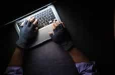 Hands with black finger gloves on them hover over a laptop keyboard in a dark setting.