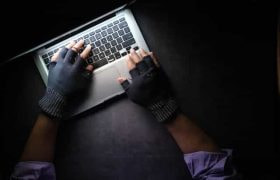 Hands with black finger gloves on them hover over a laptop keyboard in a dark setting.