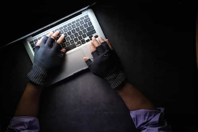 Tangan dengan sarung tangan jari hitam di atasnya melayang di atas keyboard laptop dalam pengaturan gelap.