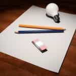 A blank sheet of paper with two pencils laying on top of it, as well as a rubber eraser and lightbulb.
