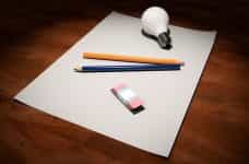 A blank sheet of paper with two pencils laying on top of it, as well as a rubber eraser and lightbulb.