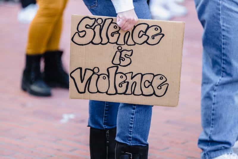 Sebuah papan karton yang dipegang di belakang kaki seorang aktivis bertuliskan, SILENCE IS VIOLENCE.