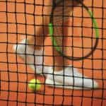 An out of focus tennis player's foot, racquet and a ball with a tennis net in the foreground.