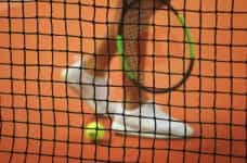 An out of focus tennis player's foot, racquet and a ball with a tennis net in the foreground.