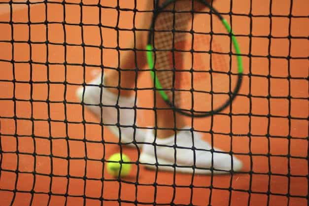 Kaki pemain tenis yang tidak fokus, raket, dan bola dengan jaring tenis di latar depan.
