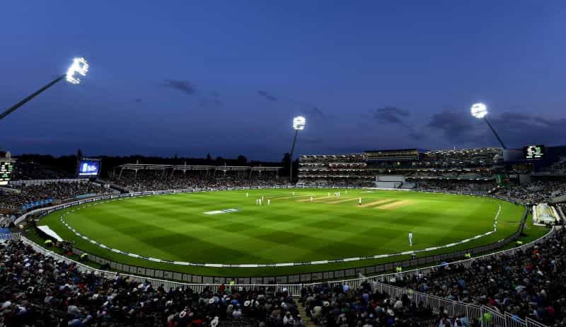 Stadion kriket yang diterangi lampu dengan permainan yang dimainkan di senja hari.