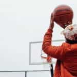 The backside of a man in an orange jacket and baseball cap throwing a basketball at a basketball hoop outside.