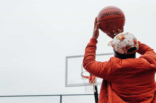Bagian belakang seorang pria berjaket oranye dan topi baseball melempar bola basket ke ring basket di luar.