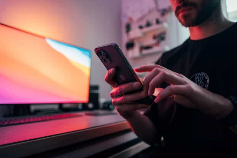 A man using his mobile phone in a pink-tinted room, with a computer monitor in the background.