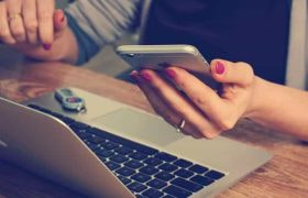 A woman sitting in front of her laptop at a desk while holding a smartphone in her left hand.