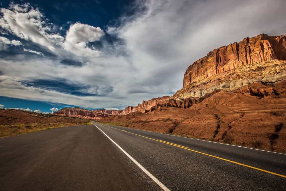 Jalan kosong yang terbuka lebar di tengah gurun pedesaan di Utah, Amerika Serikat, dikelilingi tebing merah dan pegunungan.