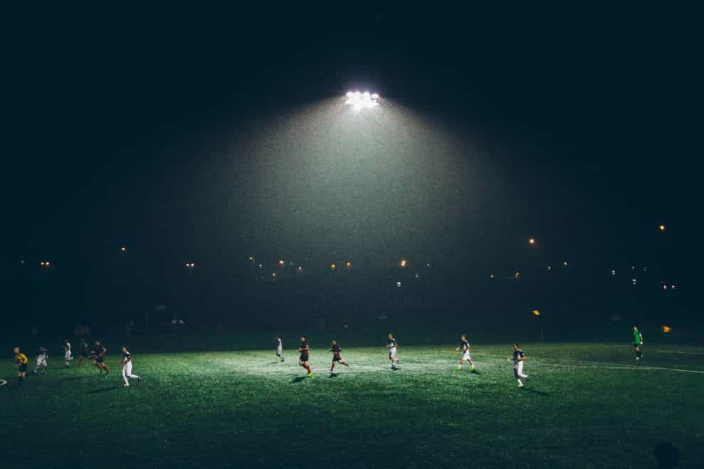 Pemain sepak bola berlari melintasi lapangan hijau di malam hari.