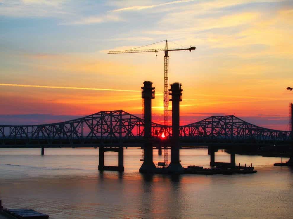 Jembatan baja besar saat matahari terbenam, dengan derek tinggi menjulang di atasnya.