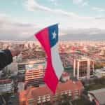 A hand holds the Chilean flag over the city of Santiago from a great height.