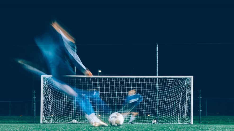 Gambar time lapse dua pemain sepak bola berseragam biru sedang menendang dan mempertahankan bola di depan gawang.