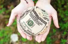 Several US dollar bills rolled up into a snowball-sized ball resting in the open palms of a pair of outstretched hands.