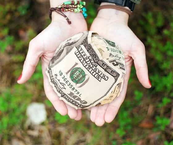 Beberapa lembar uang dolar AS digulung menjadi bola seukuran bola salju yang diletakkan di telapak tangan terbuka dari sepasang tangan yang terulur.