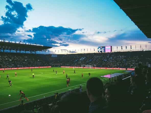Stadion sepak bola yang penuh sesak di bawah lampu sorot dengan pertandingan langsung sedang berlangsung.