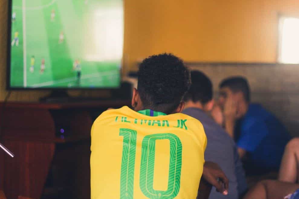 Seorang pria duduk di bar menonton pertandingan sepak bola di TV sambil mengenakan jersey kuning Neymar Jr. Brasil.