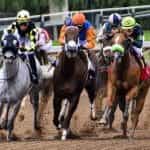 Five jockeys in different colored outfits racing their horses on a race track.