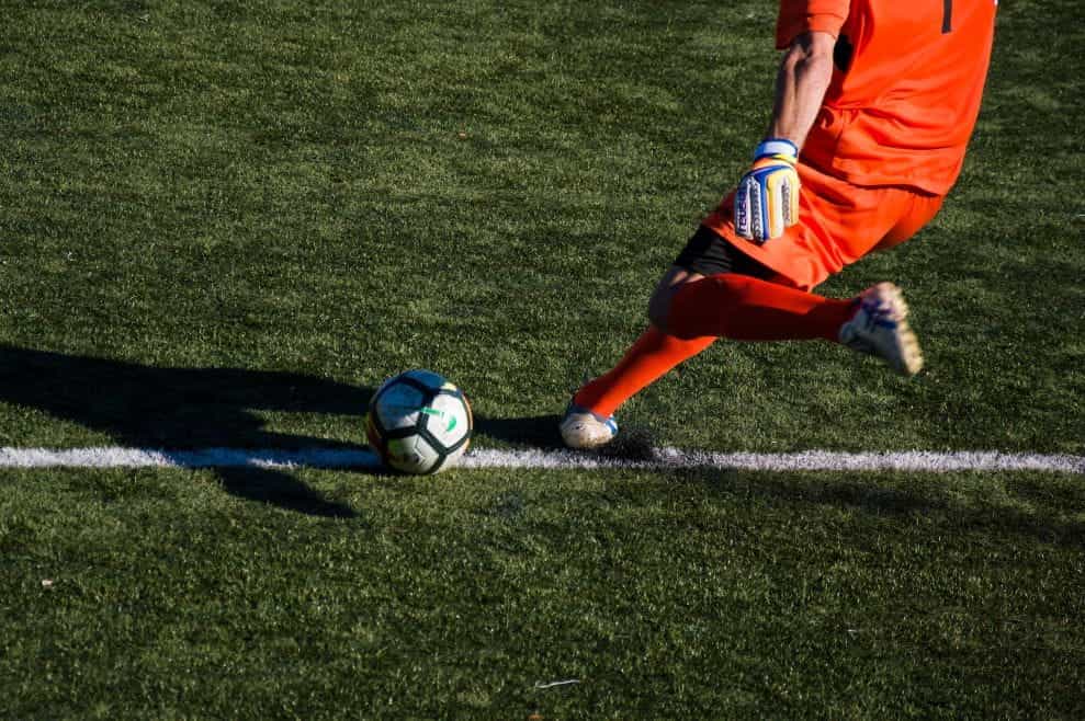 Seorang pemain sepak bola berseragam oranye bersiap untuk menendang bola di lapangan hijau.