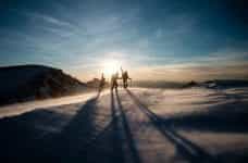 Three people with skiing gear walk towards the summit of a snow covered mountain as the sun sets.