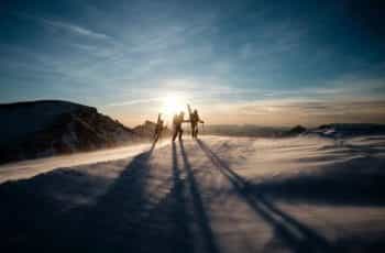 Three people with skiing gear walk towards the summit of a snow covered mountain as the sun sets.