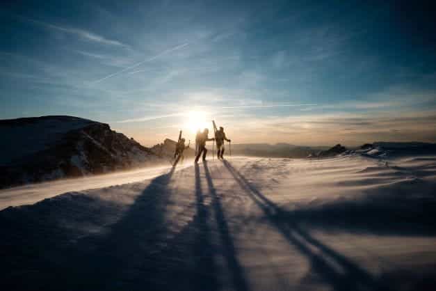 Tiga orang dengan peralatan ski berjalan menuju puncak gunung yang tertutup salju saat matahari terbenam.