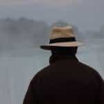 The back of a man wearing a brown jacket and a beige fedora and looking into the mist.
