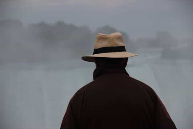 Bagian belakang seorang pria yang mengenakan jaket coklat dan topi fedora krem ​​sedang melihat ke dalam kabut.