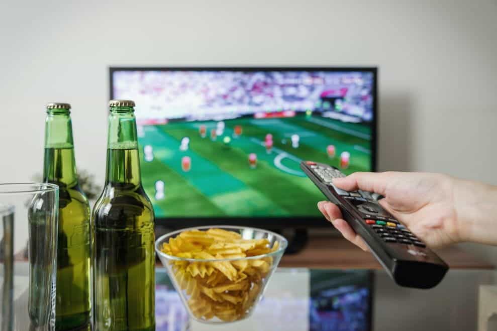 Sebuah tangan memegang remote menunjuk ke TV yang menayangkan pertandingan sepak bola, dengan bir dan keripik.