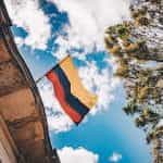 The Colombian flag waves between a building, trees, and a blue sky.