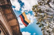 The Colombian flag waves between a building, trees, and a blue sky.