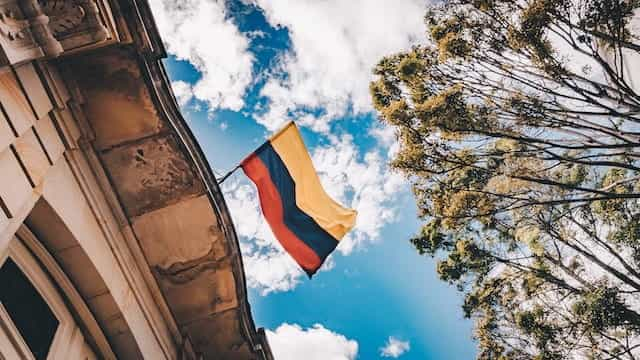 Bendera Kolombia melambai di antara gedung, pepohonan, dan langit biru.