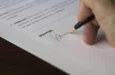A person’s hand holding a pen and signing a written contract on paper with an official signature.