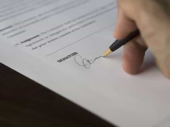 Tangan seseorang memegang pena dan menandatangani kontrak tertulis di atas kertas dengan tanda tangan resmi.