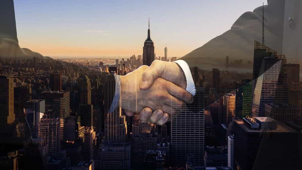 Dua sosok tembus pandang berjabat tangan di depan latar belakang yang memperlihatkan cakrawala tengah kota Manhattan di New York City.